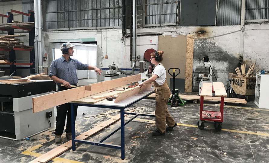 Dans un atelier, un homme et une femme sont face à face autour d’une table sur laquelle sont posées deux planches de bois