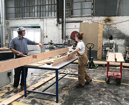 Dans un atelier, un homme et une femme sont face à face autour d’une table sur laquelle sont posées deux planches de bois