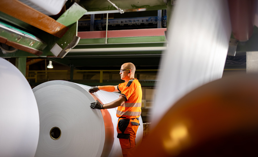 Une photographie avec la vue intérieure d'une usine, un immense rouleau de papier et un ouvrier.