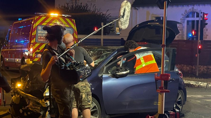 Une vidéaste et un preneur de son filment une voiture accidentée et son chauffeur secouru par un pompier